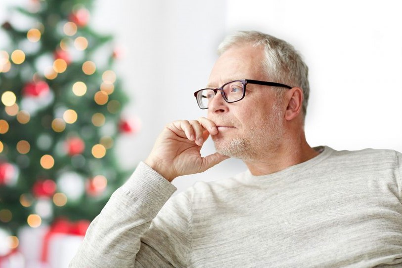 A man is sitting alone on a couch, with a Christmas tree in the background. He is alone for the holidays.