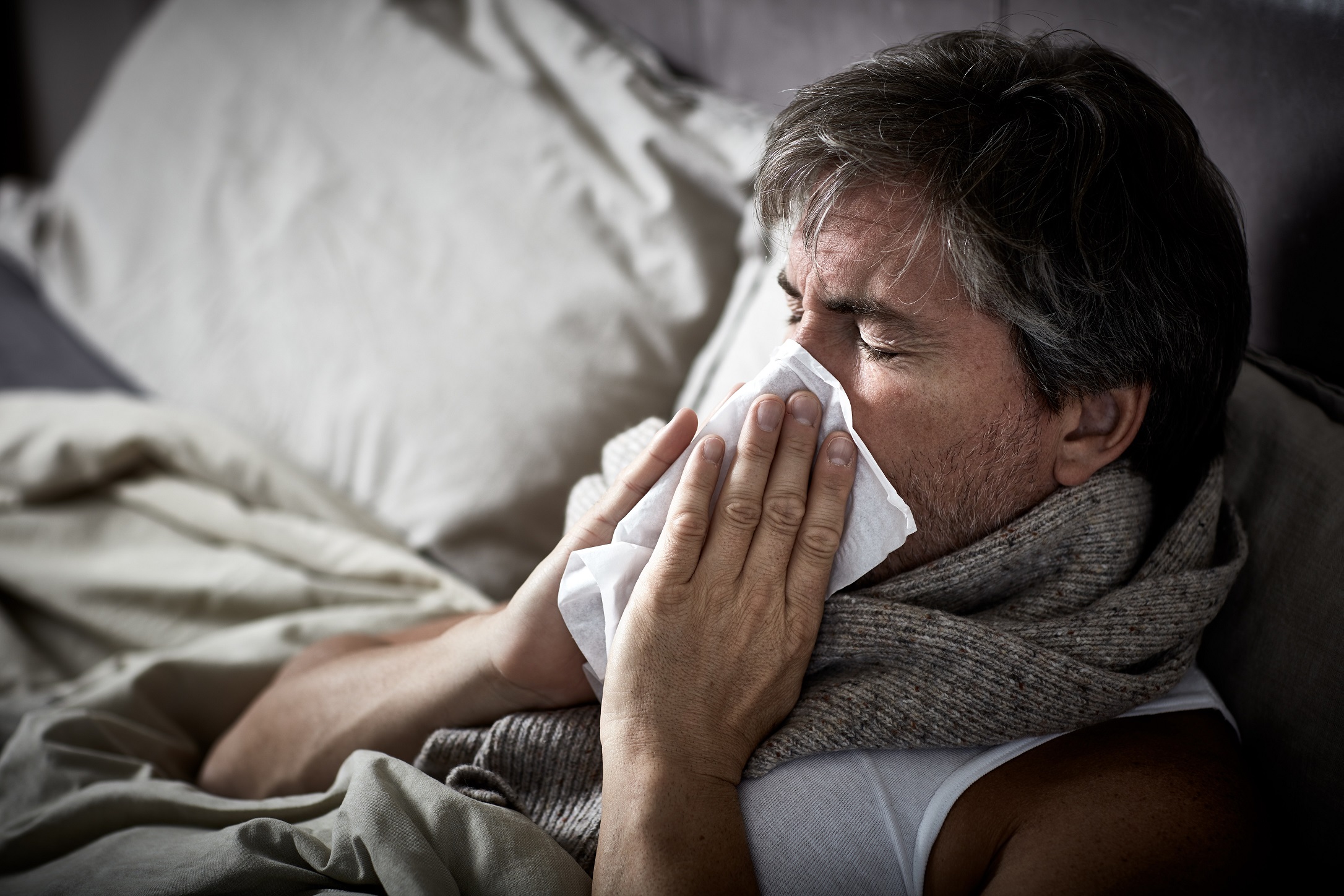 A middle-aged man is in bed with the flu, sneezing into a tissue.