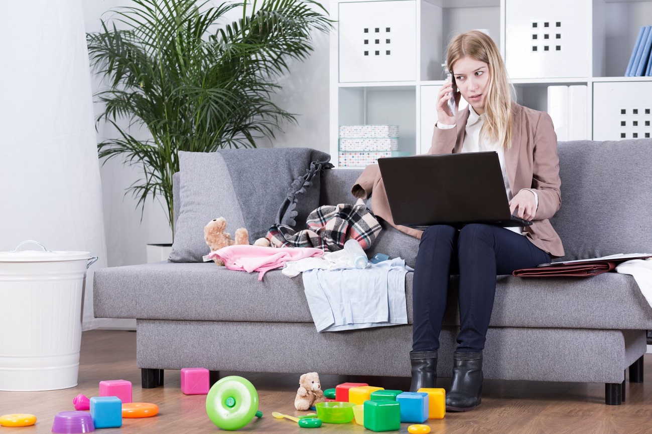 A woman working from home, on her couch, with a laptop on her lap and toys scattered at her feet and on the couch.
