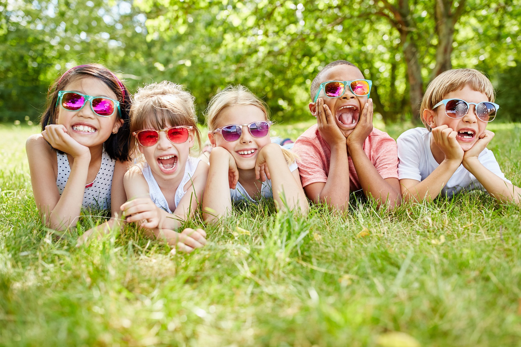 Five smiling boys and girls crouched on the ground with their hands under their chins, wearing colorful sunglasses