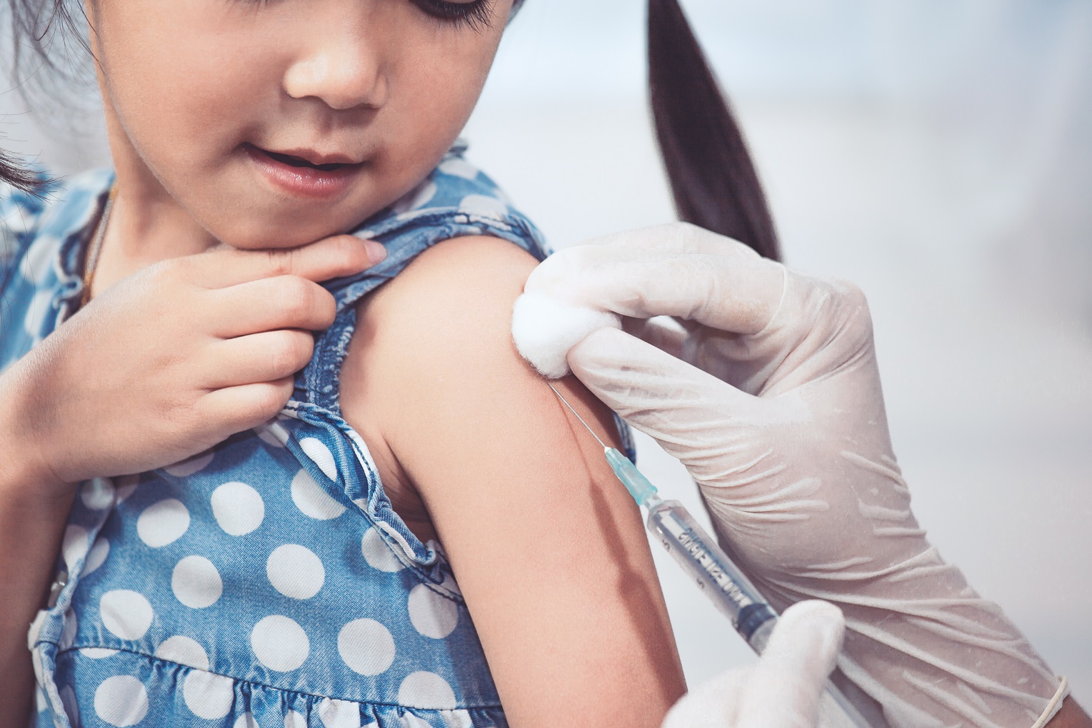 A young girl wearing a blue and white polka dotted top looks at the hand of her doctor who is applying alcohol to her shoulder before giving her an immunization.