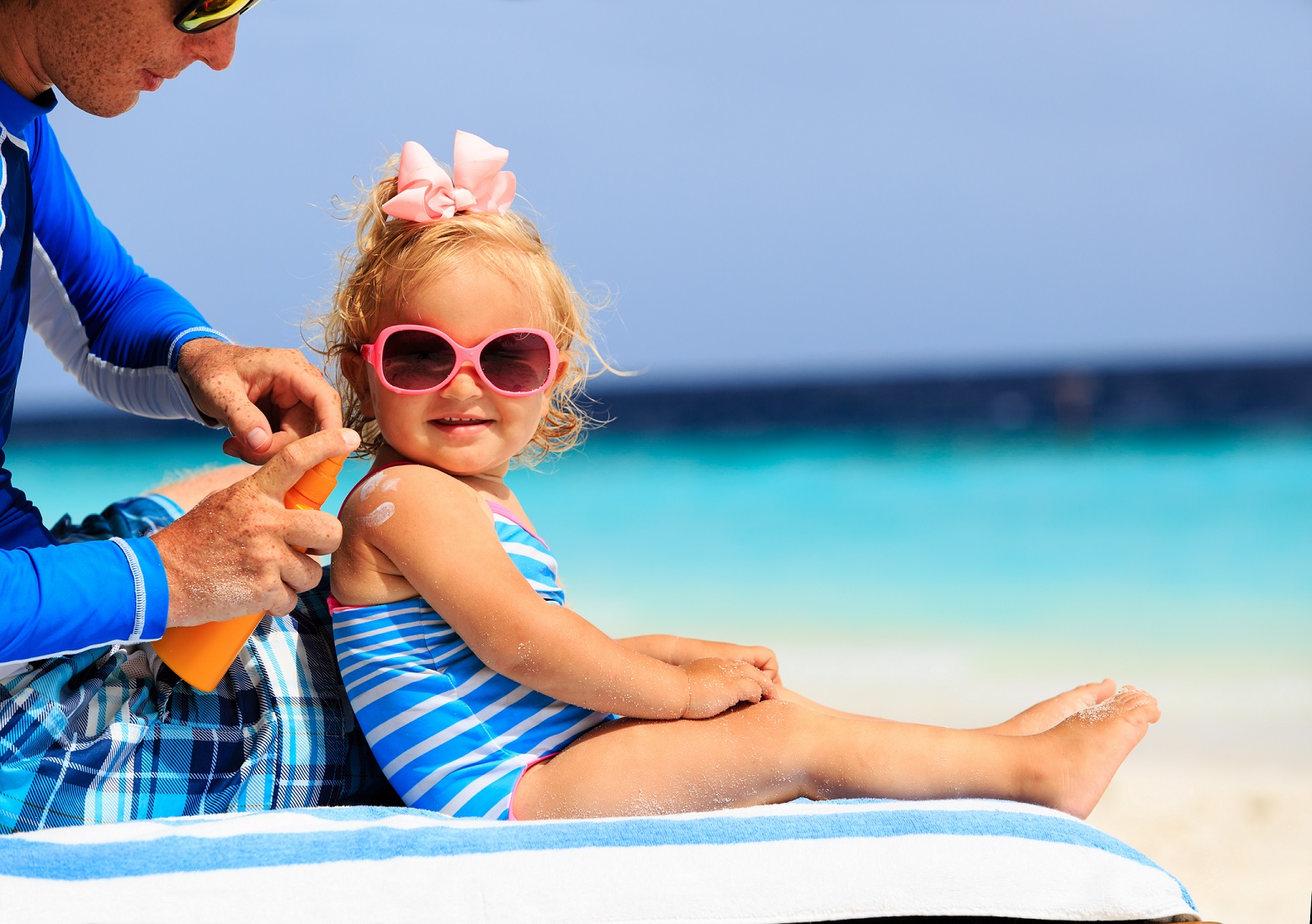 A photo of a blonde little girl wearing sunglasses, with her father who is applying sunscreen.