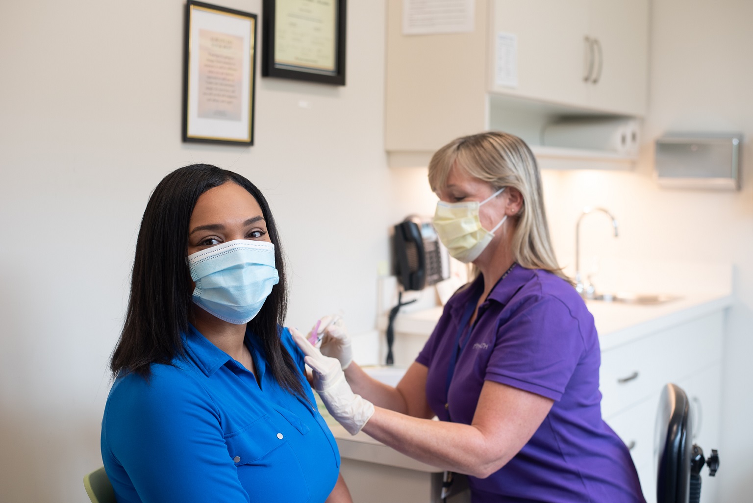 A brunette wearing a blue shirt and surgical mask gets a flu shot from a blonde RVNAhealth nurse who is wearing a purple shirt and a face mask