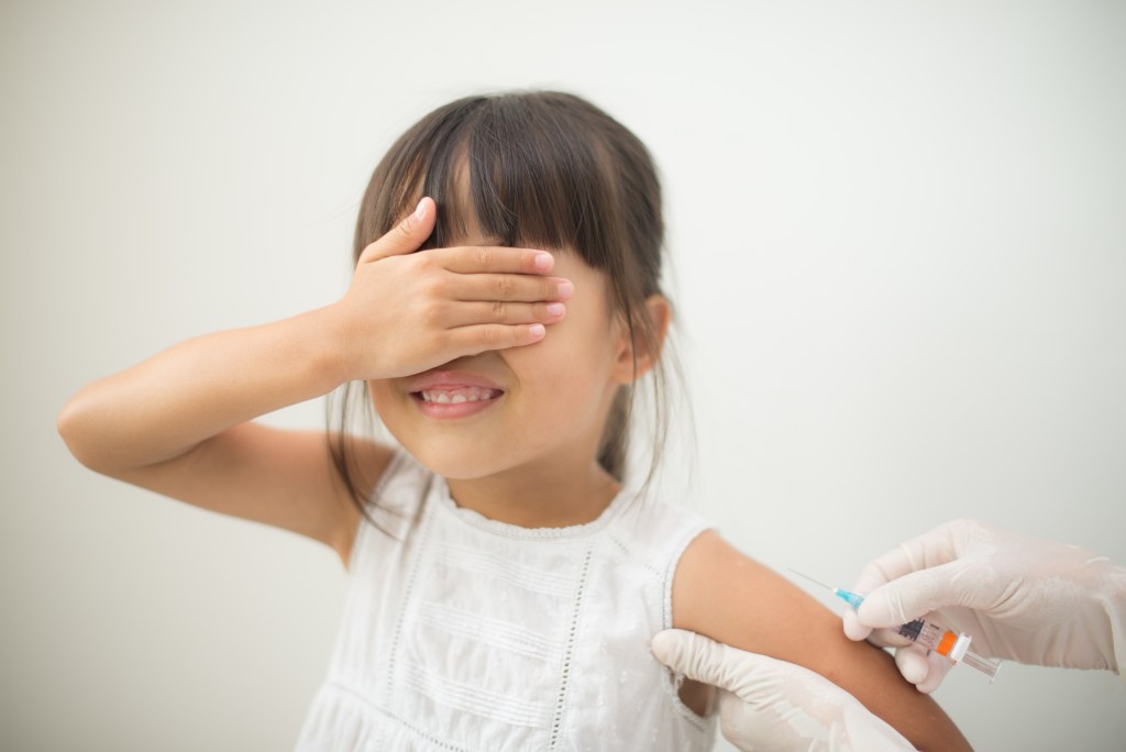 A young girl with brown hair covers her eyes while getting a shot.