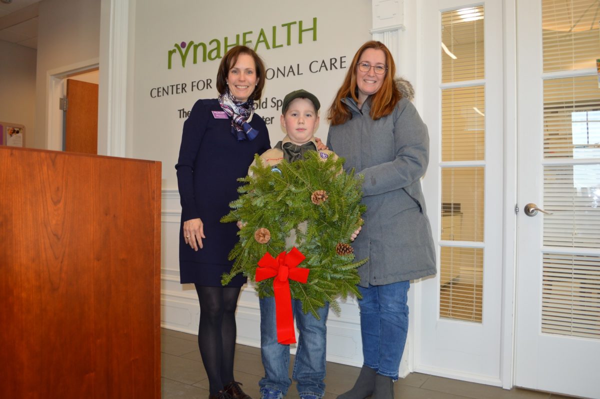A young boy wearing a cub scout uniform holds a wreath that he is donating. He is flanked by his mother and RVNA Director of Philanthropy Mary Jean Heller