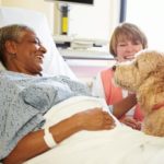 A woman in a hospital bed and a volunteer pet companion laugh at an affectionate portuguese water dog who is sitting by the bed