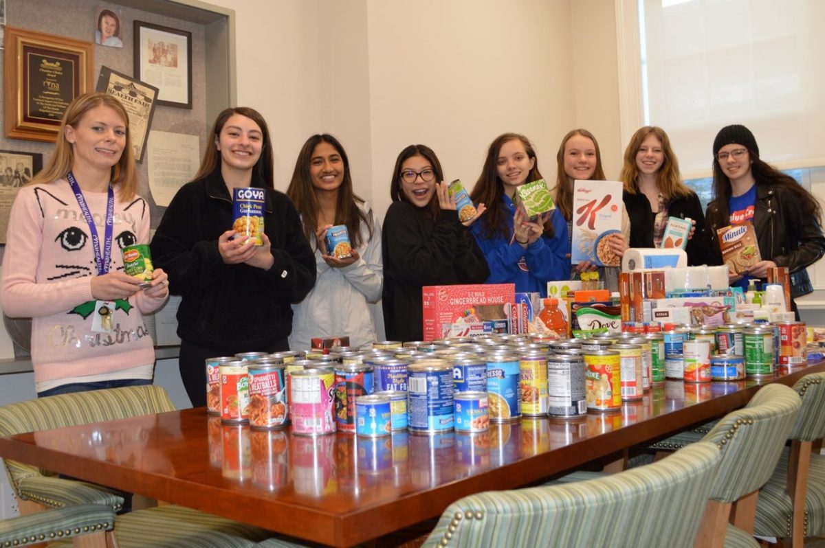 Six teenage girls stand behind a table stacked with food pantry donations that they collected as youth volunteers.. Standing next to them is RVNAhealth programs coordinator Katelyn Scribner.