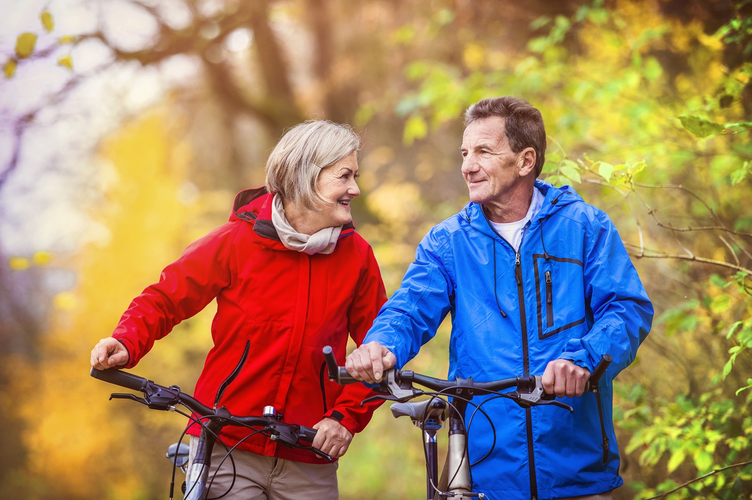 A smiling woman wearing a red windbreaker and a smiling man with a red windbreaker walk with their bicycles