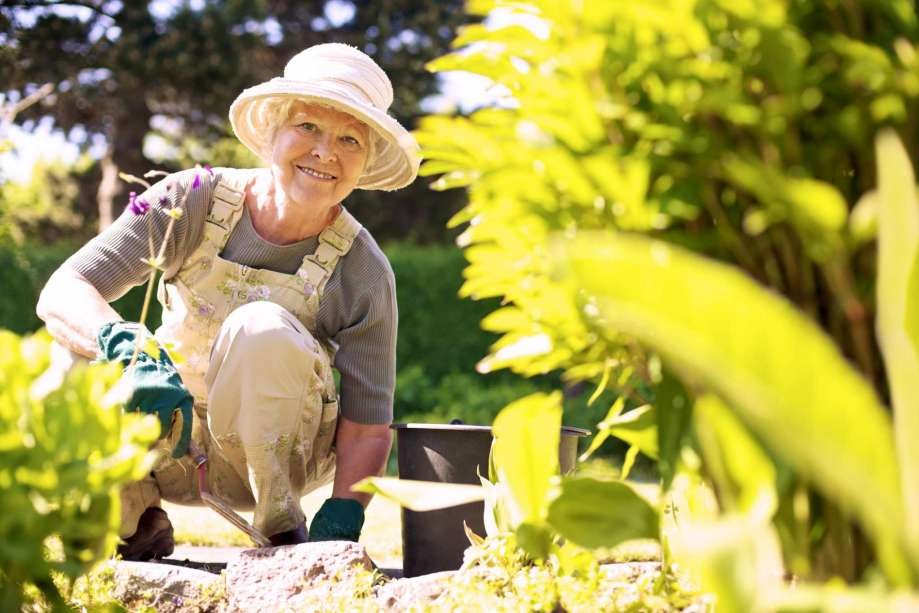 A smiling middle aged woman wearing a hat and overalls kneels in her garden to work