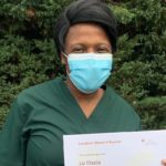 A woman wearing green scrubs and a blue surgical mask holds up her Above and Beyond certificate